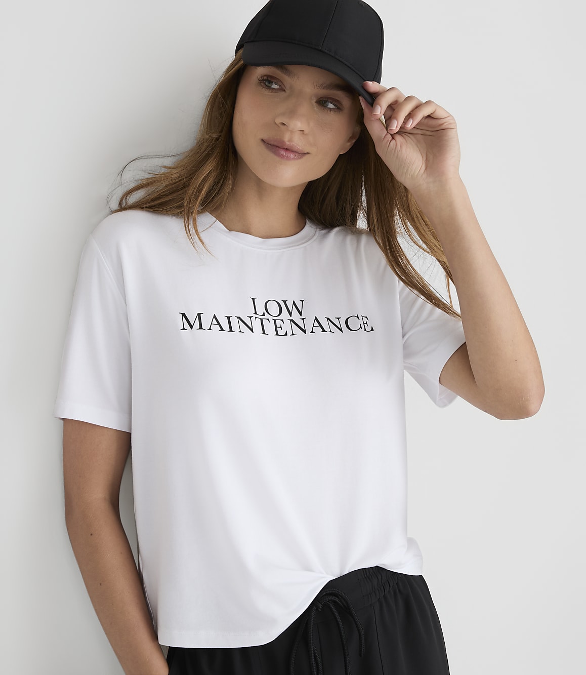 A woman in a white \"LOW MAINTENANCE\" t-shirt and black baseball cap poses against a plain white background.