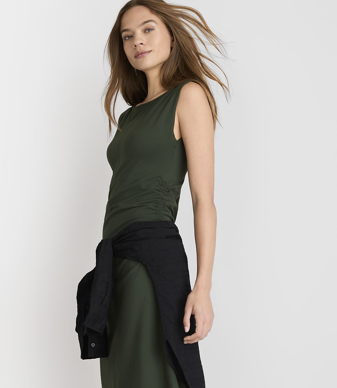 A woman in a green dress with a black sweater tied around her waist stands against a plain white background.