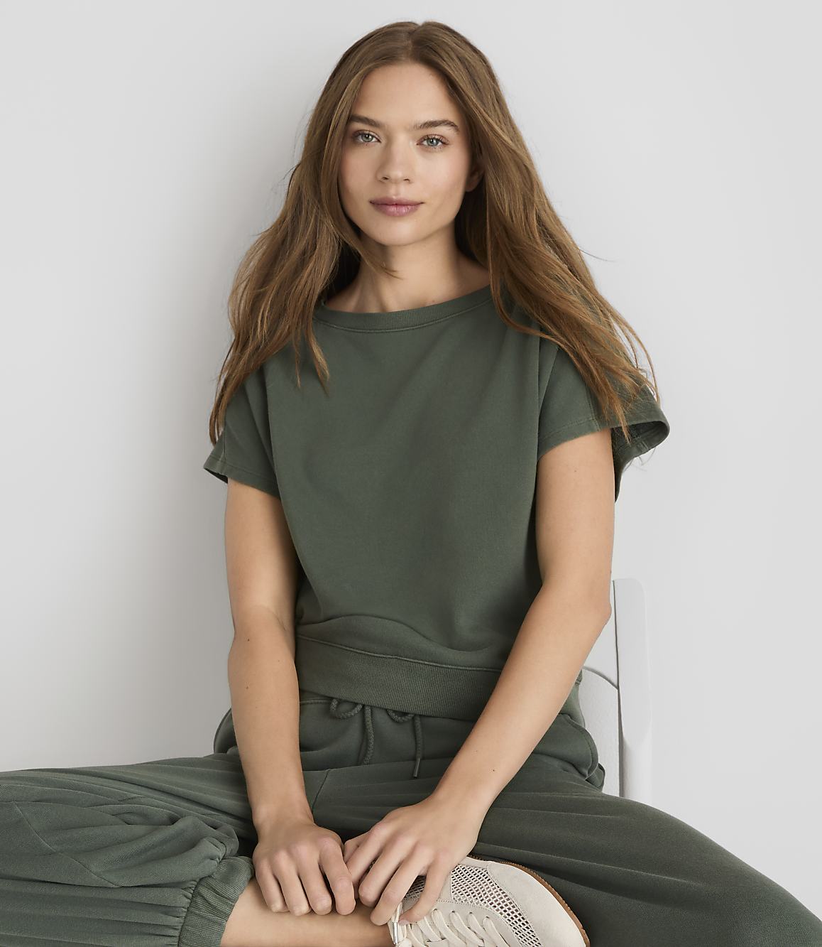 A young woman with long, wavy brown hair, wearing a green t-shirt and pants, sits on a white chair against a plain white background.