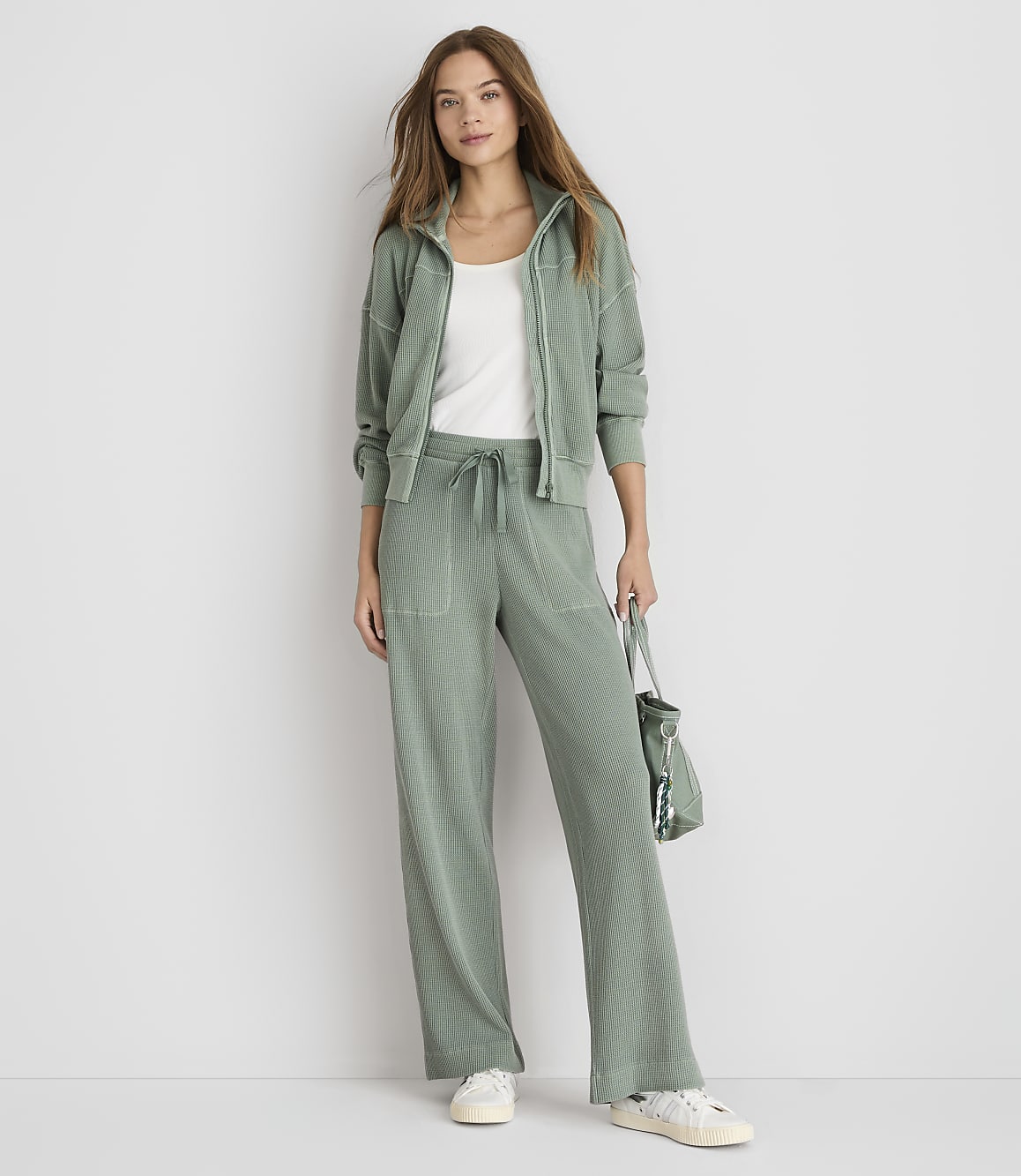A woman in a sage green loungewear set stands against a plain white background, holding a matching bag.