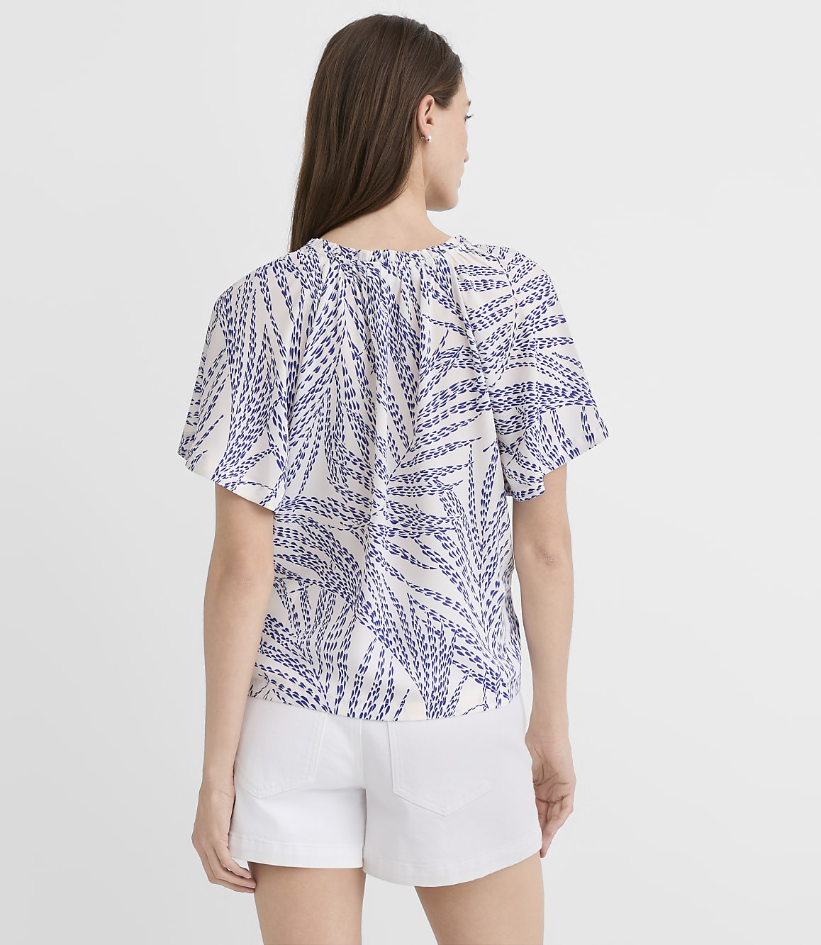 A woman in a white and blue patterned top and white shorts stands against a plain white background.