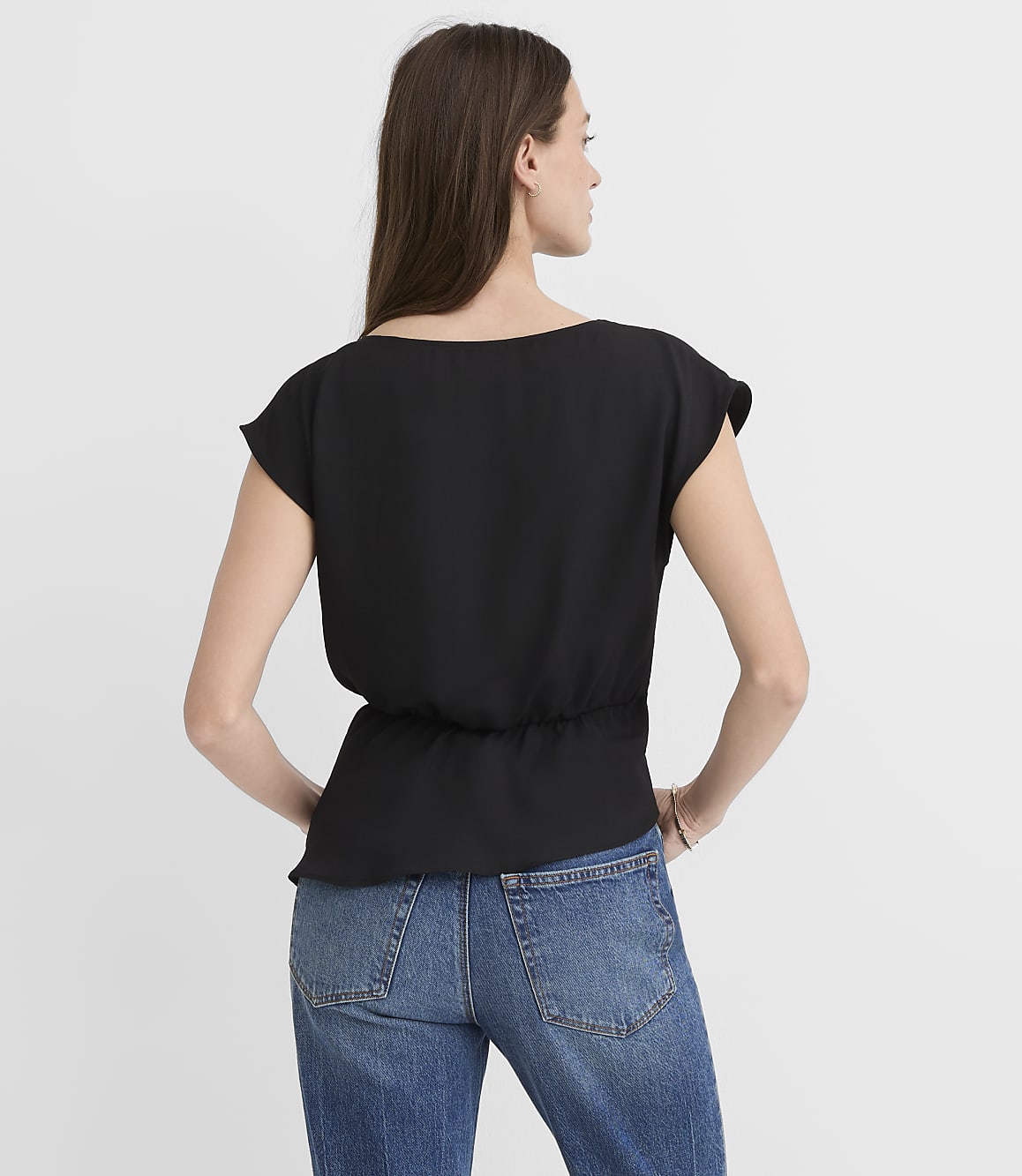 A woman in a black top and blue jeans stands against a plain white background.