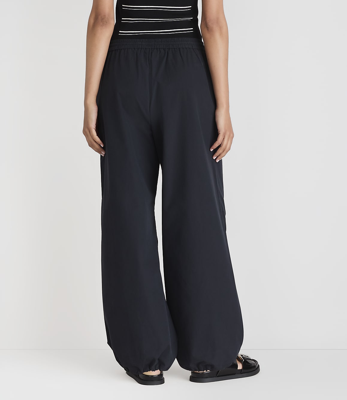 A person wearing dark, wide-leg pants and sandals stands against a plain white background.