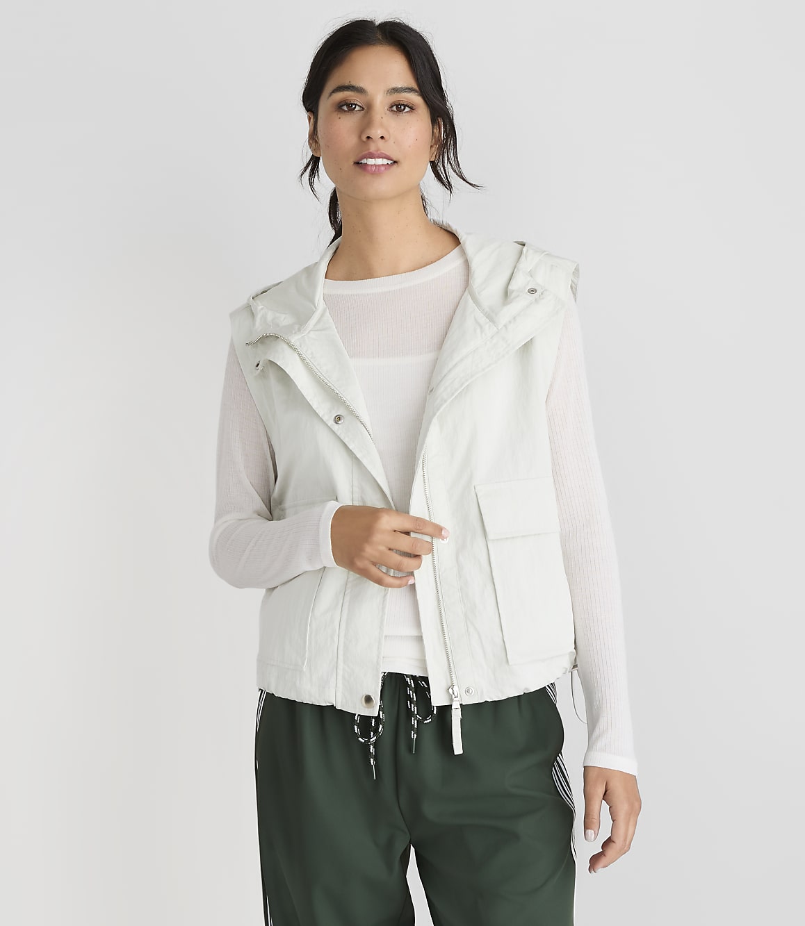 A woman in a light-colored hooded vest and green pants stands against a plain white background.