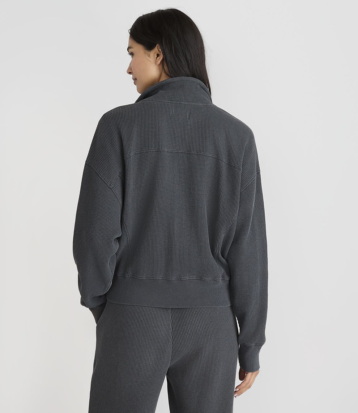 A woman in a dark grey ribbed quarter-zip sweatshirt and matching pants stands against a plain white background.