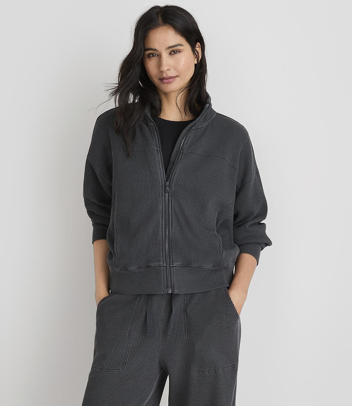A woman in a dark grey zip-up jacket and matching pants stands against a plain white background.