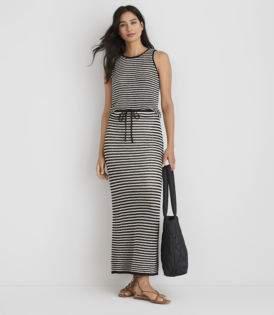 A woman in a striped maxi dress and sandals stands against a plain white background, holding a black quilted tote bag.