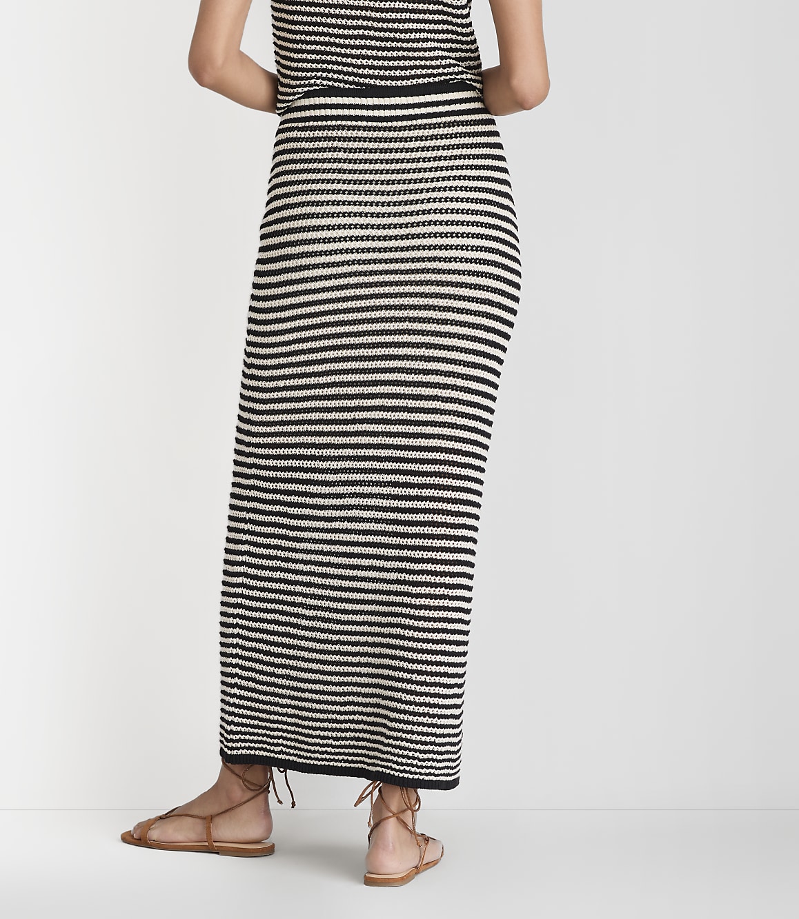A person wearing a black and white striped maxi skirt and brown sandals stands against a plain white background.