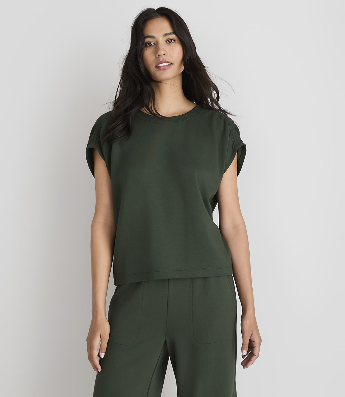A woman in a dark green top and pants stands against a plain white background.
