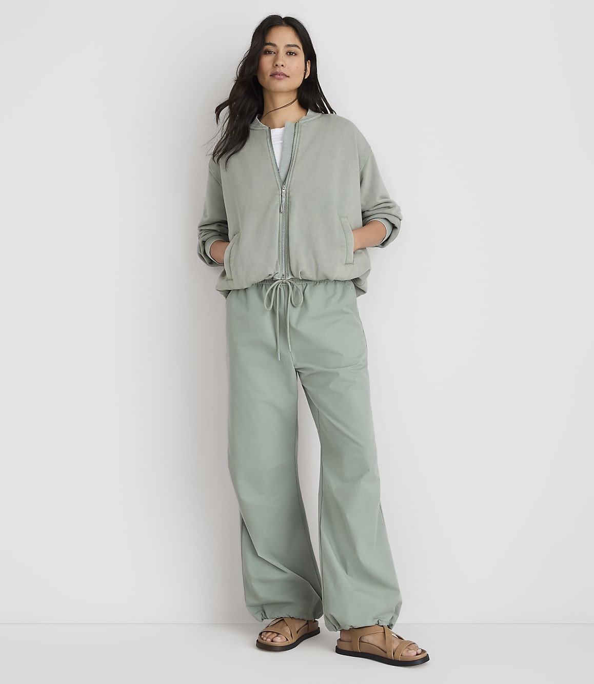 A woman stands in the foreground wearing a sage green bomber jacket and matching wide-leg pants, against a plain white background.