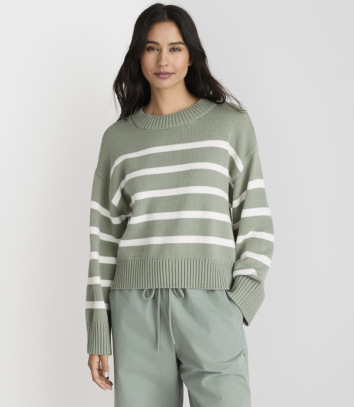 A woman in a green and white striped sweater and light green pants stands against a plain gray background.