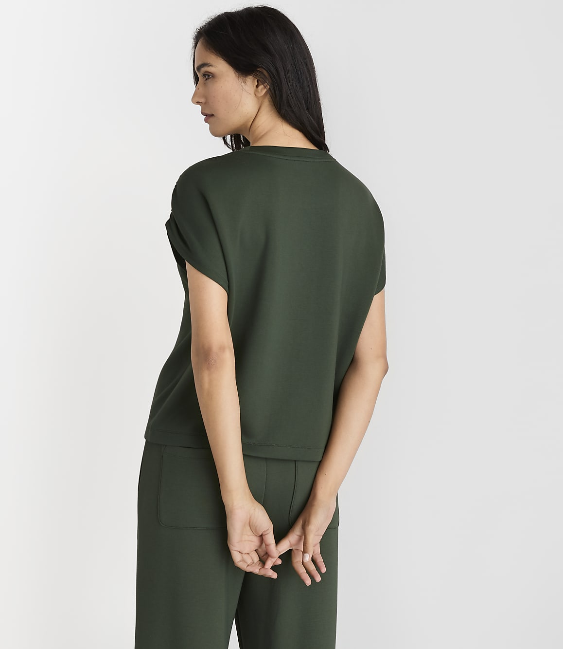 A woman in a dark green top and pants stands with her back to the viewer against a plain white background.