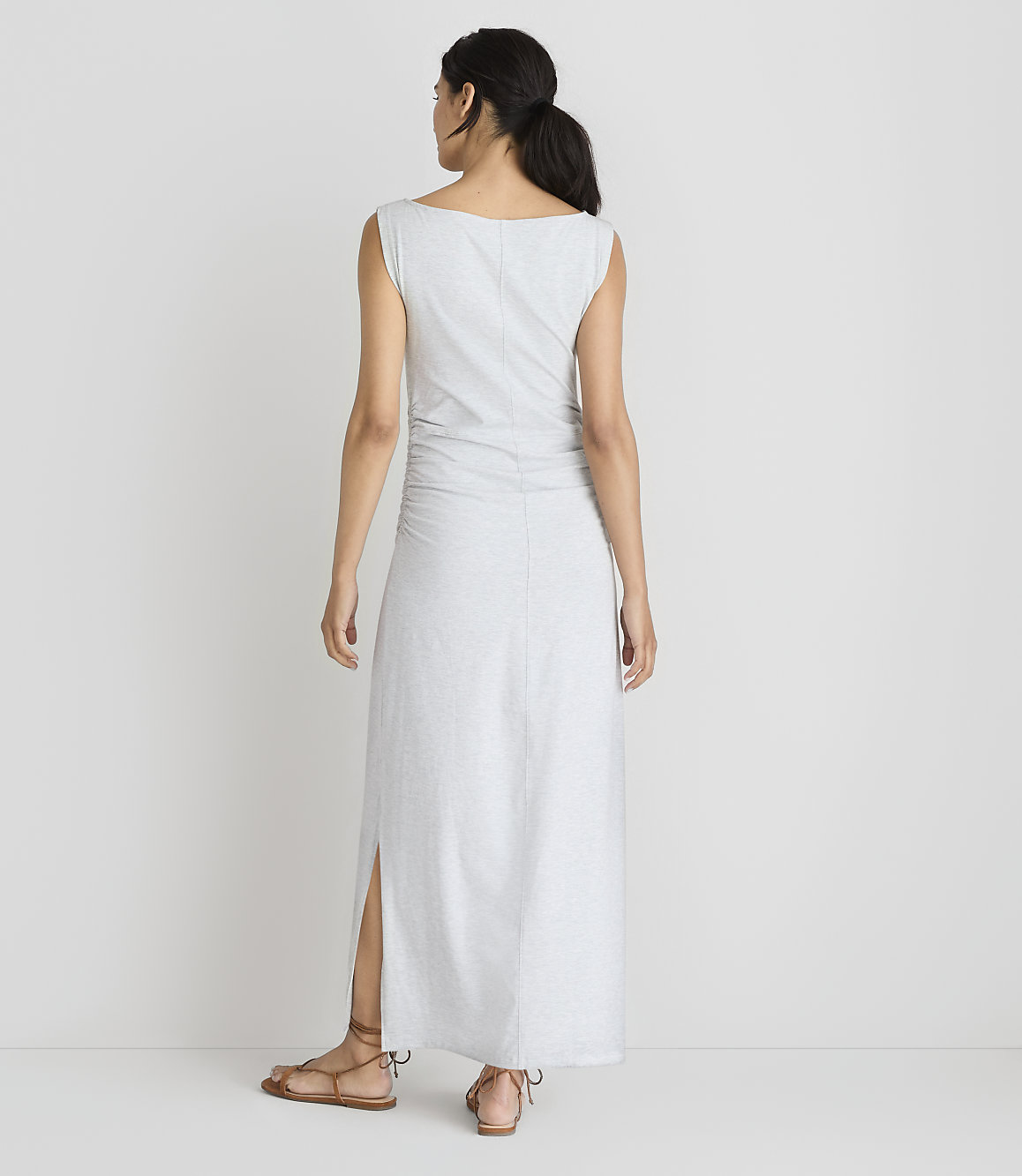 A woman in a light gray maxi dress with ruched detailing and a side slit stands with her back to the camera against a plain white background.