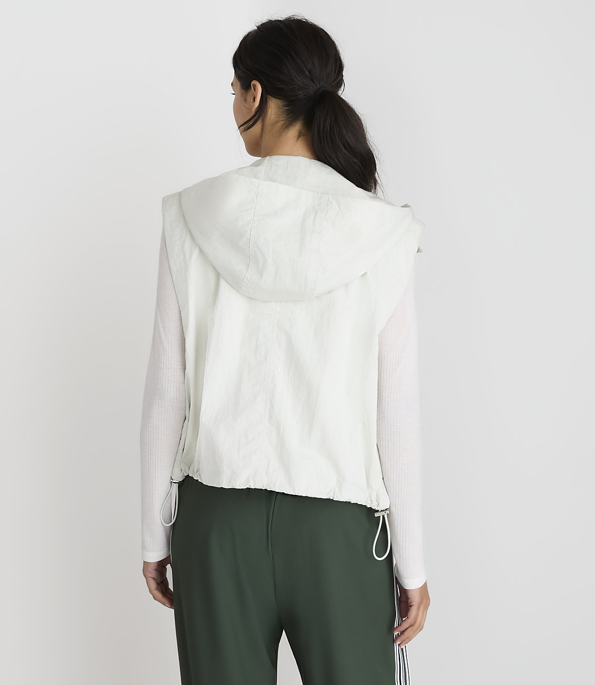 A woman in a white hooded vest and green pants stands against a plain white background.