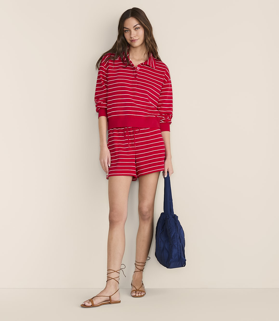 A woman in a red and white striped polo shirt and shorts stands against a plain, light-colored background, holding a blue bag.