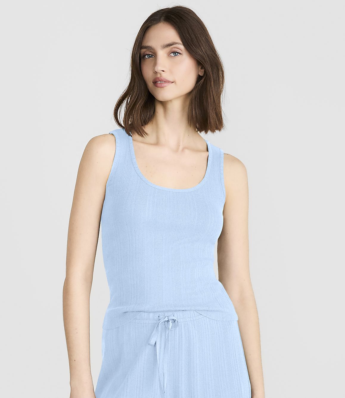 A woman in a light blue ribbed tank top and matching pants stands against a plain white background.