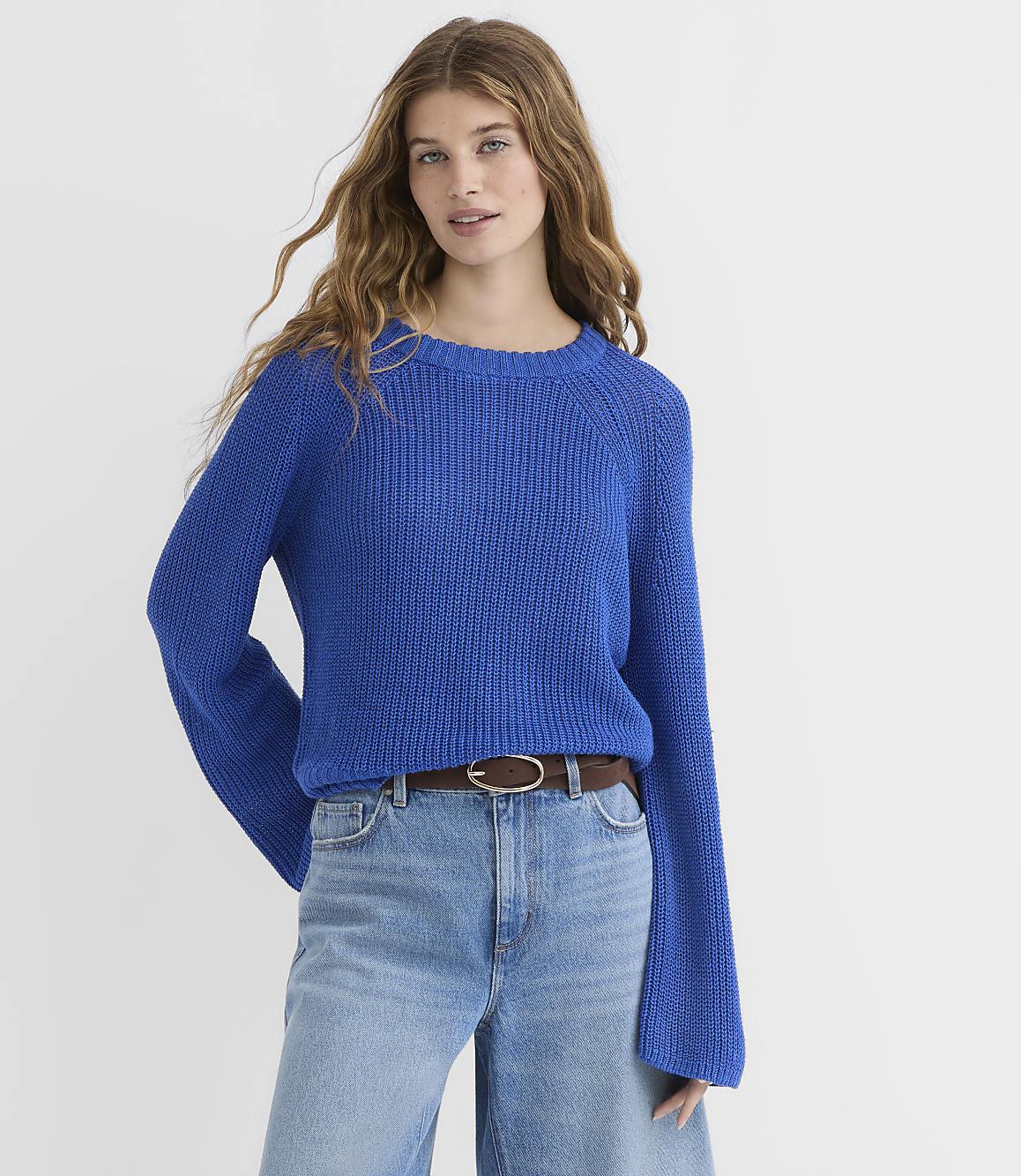 A young woman in a blue sweater and jeans stands against a plain white background.