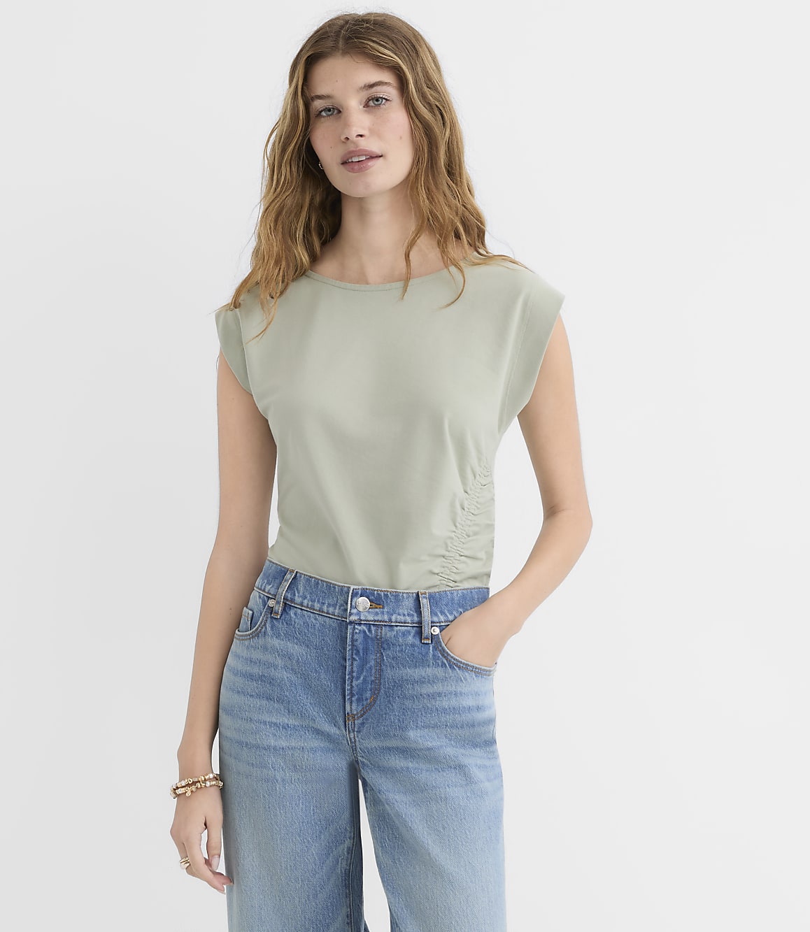 A woman in a light green top and blue jeans stands against a plain white background.