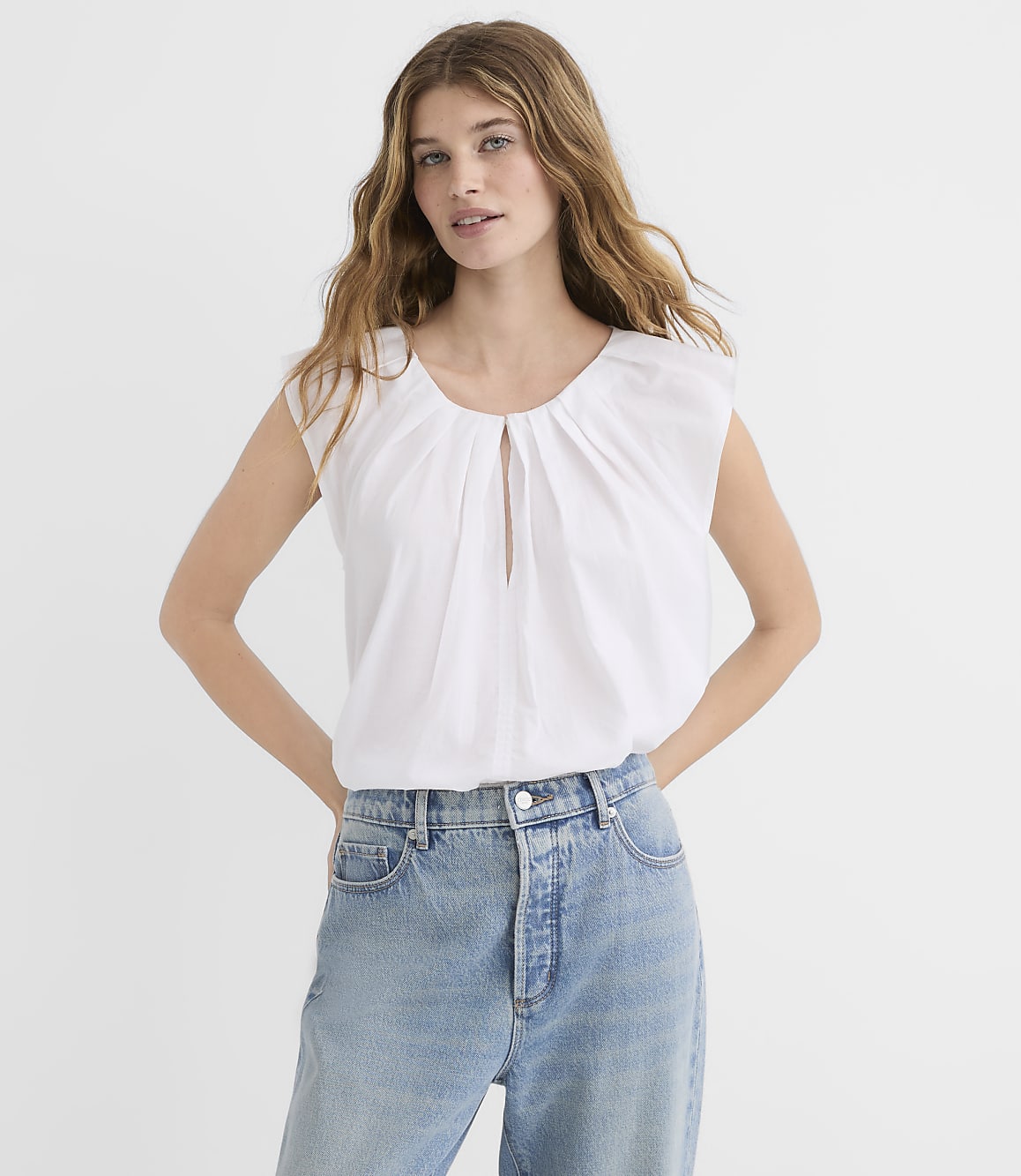 A woman in a white top and blue jeans stands against a plain white background.