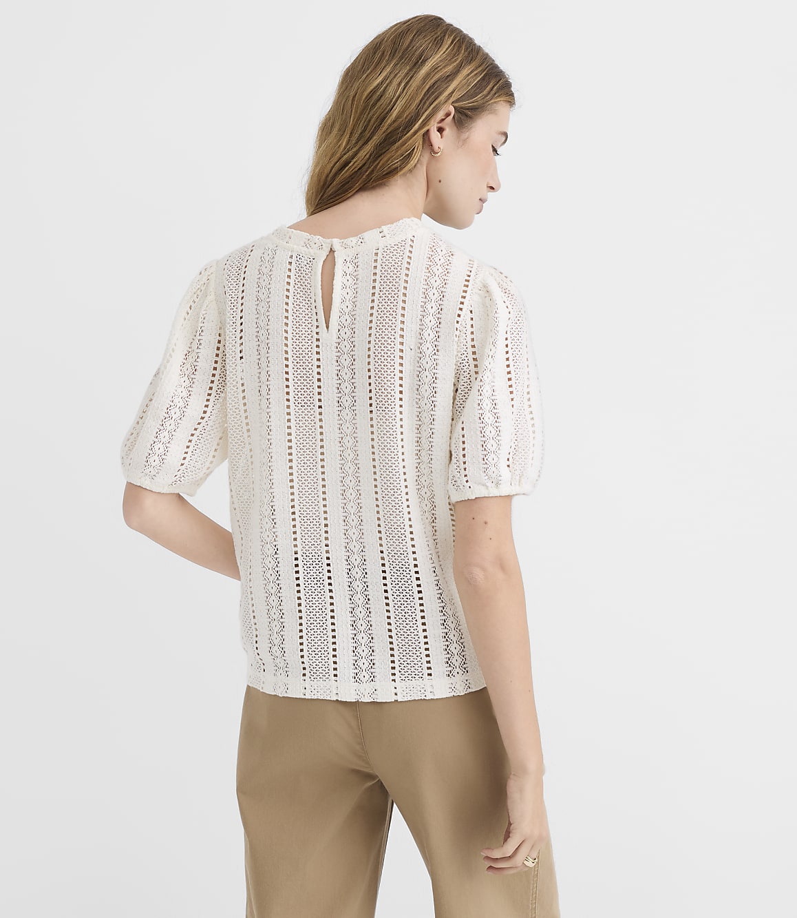 A woman in a white eyelet top and tan pants stands against a plain white background.
