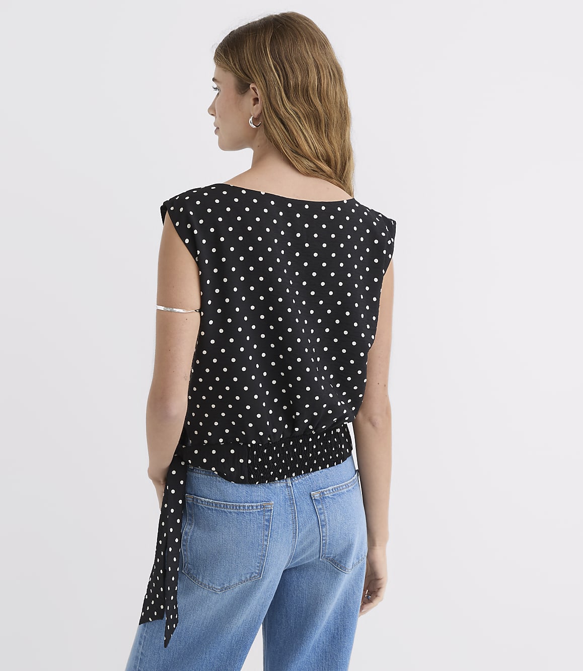A woman in a black polka dot top and blue jeans stands against a plain white background.