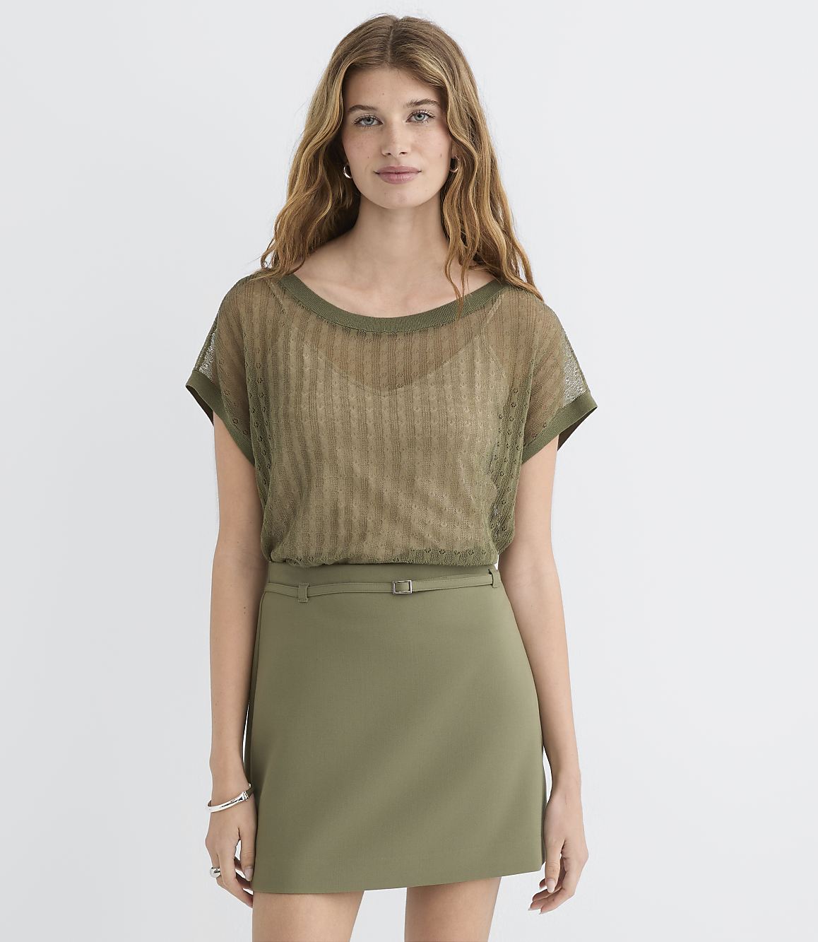A woman in a sheer olive green top and olive green skirt stands against a plain white background.