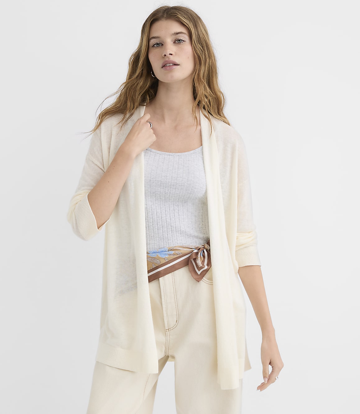 A woman in a cream cardigan and light-colored pants stands against a plain white background.