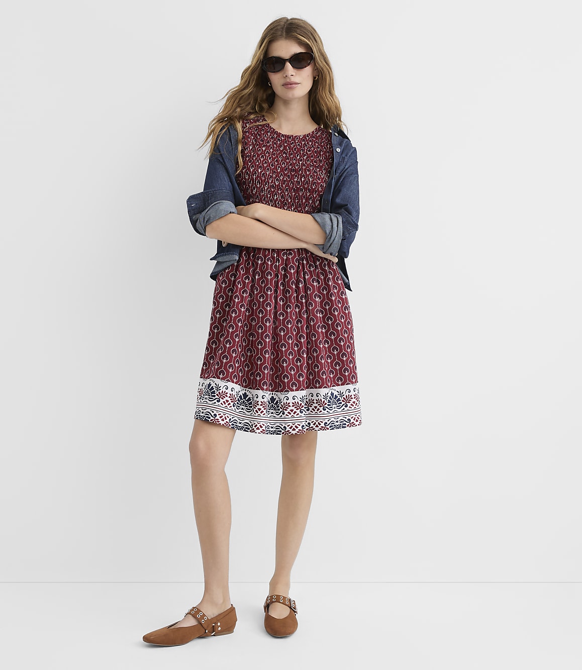 A woman in a patterned dress and denim jacket stands with her arms crossed against a plain white background.