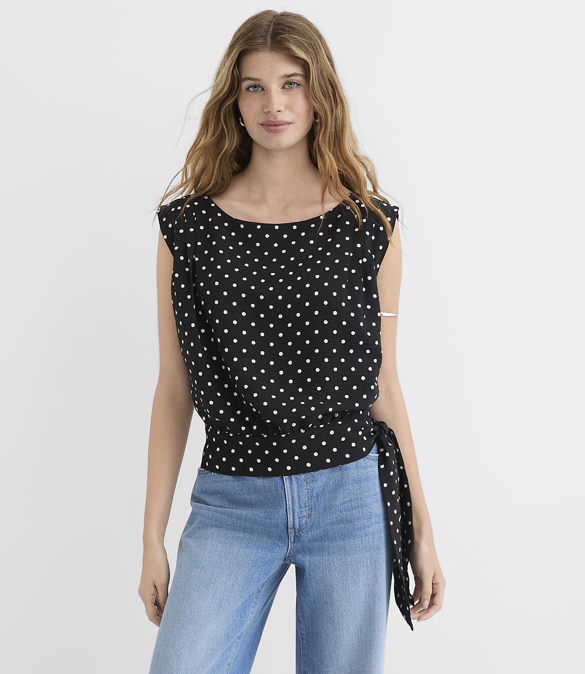 A woman in a black polka dot top and blue jeans stands against a plain white background.