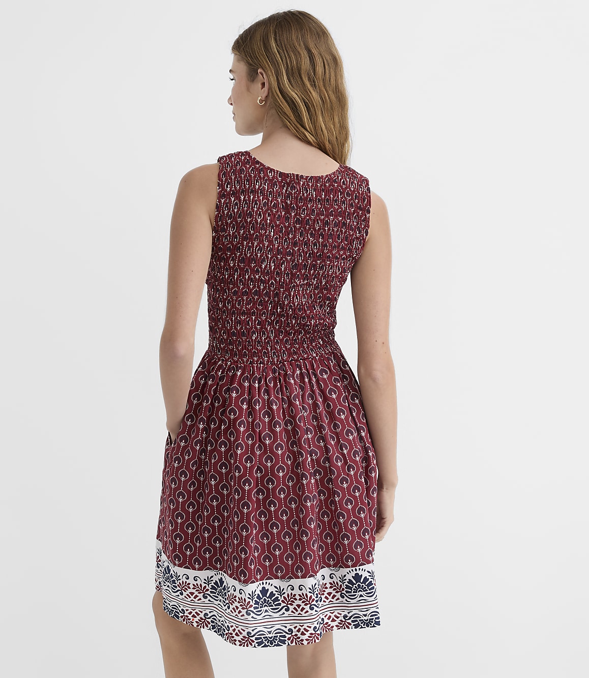A woman in a patterned maroon dress with smocked detailing stands against a plain white background.