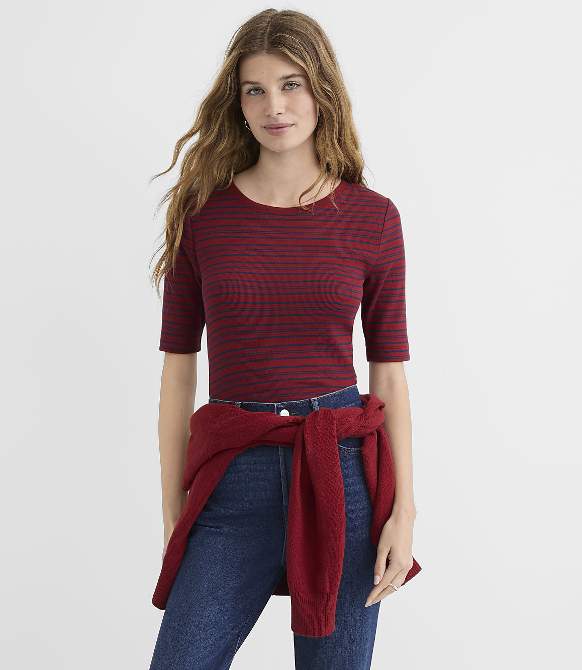 A woman in a striped shirt and jeans stands against a plain white background, with a red sweater tied around her waist.