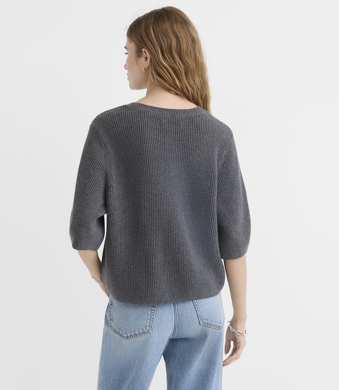 A woman in a grey sweater and jeans stands against a plain grey background.