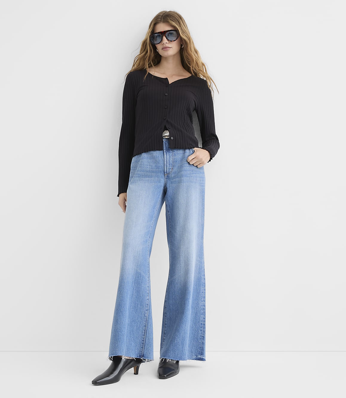 A woman in a black cardigan and wide-leg jeans stands against a plain white background.