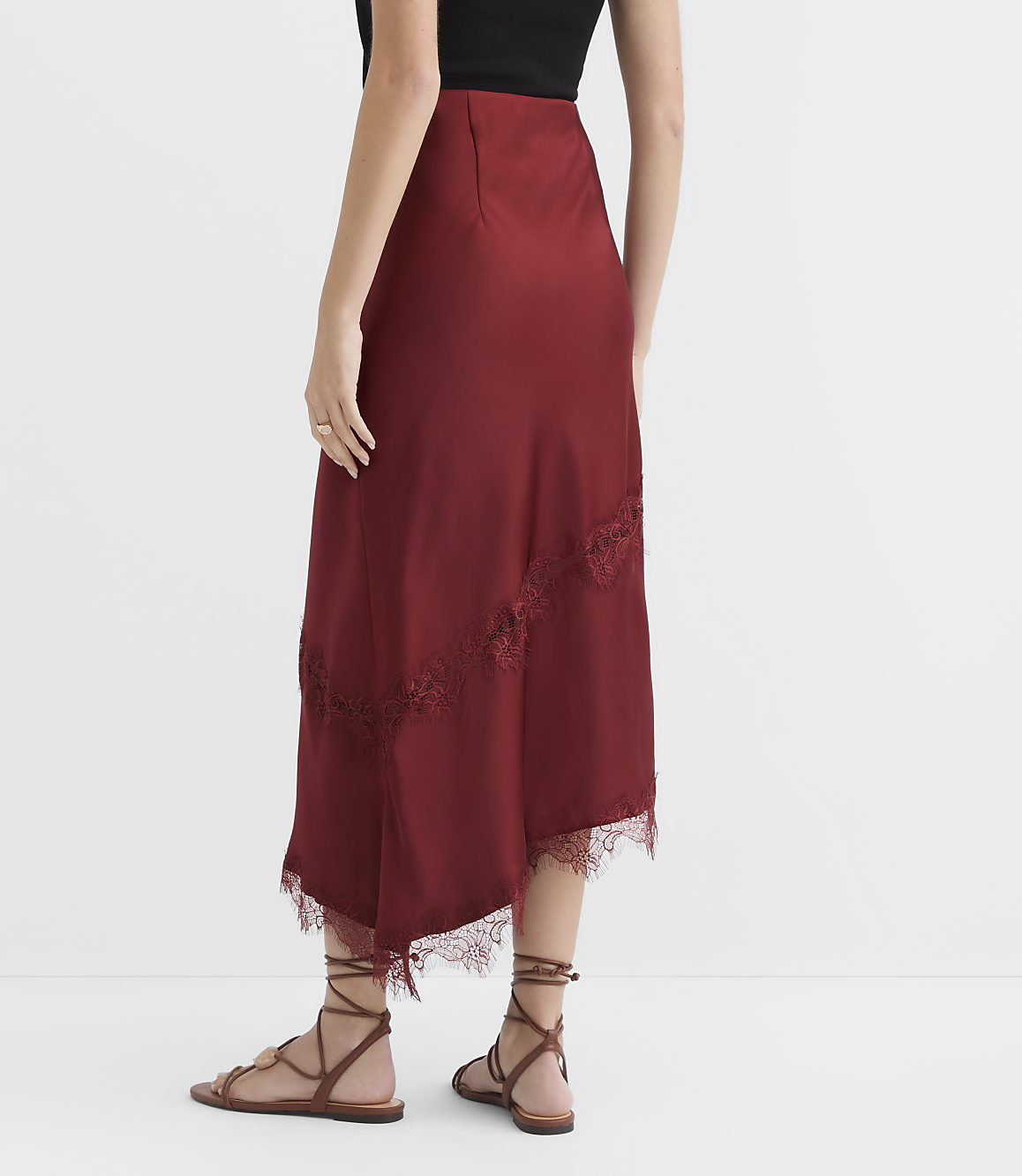 A woman in a burgundy satin skirt with lace trim stands on a white floor, with a plain white background behind her.
