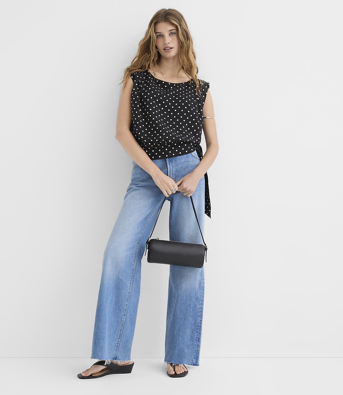 A woman in a polka dot top and wide-leg jeans stands against a plain white background, holding a black cylindrical purse.