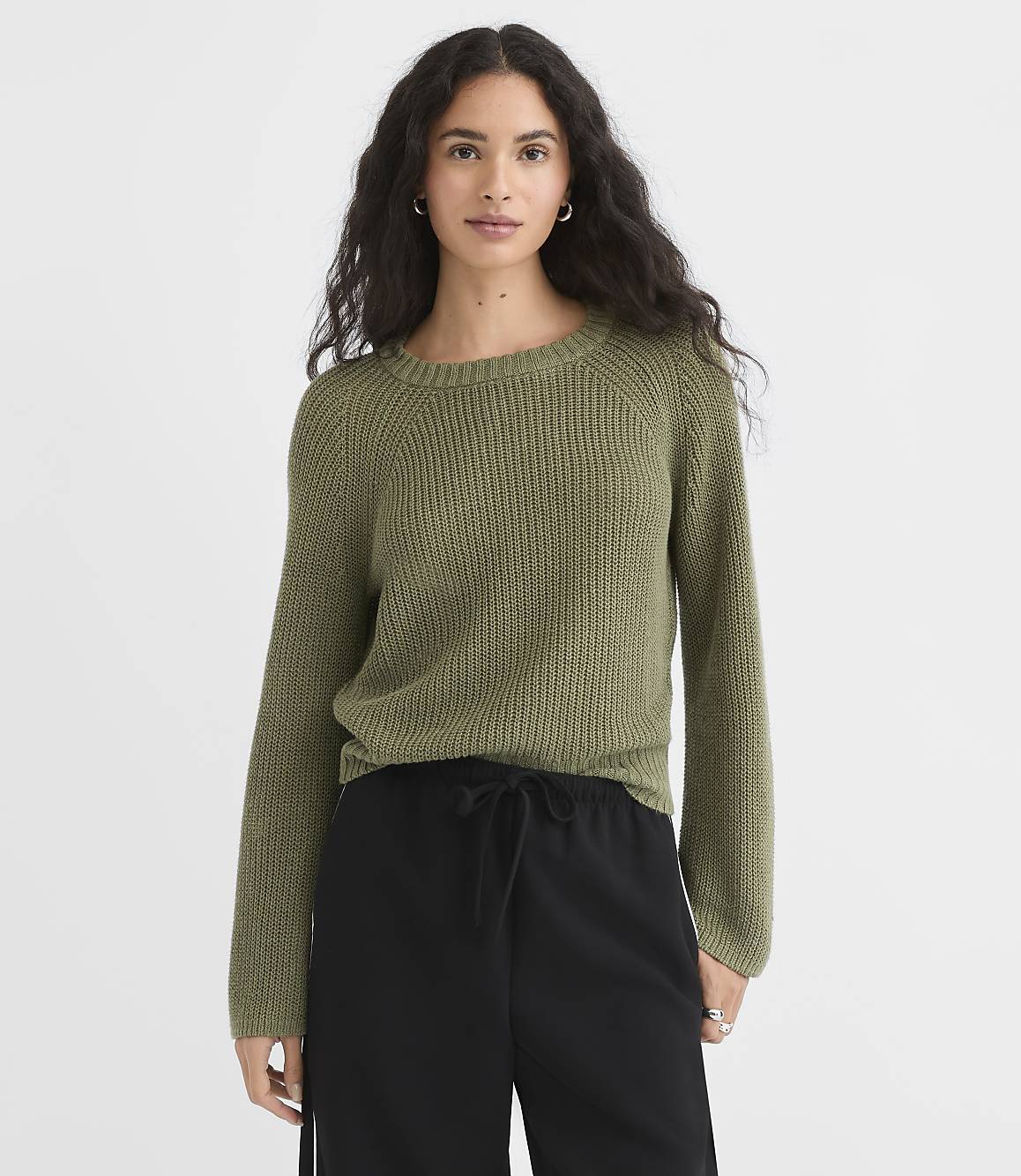 A woman in a green sweater and black pants stands against a plain white background.