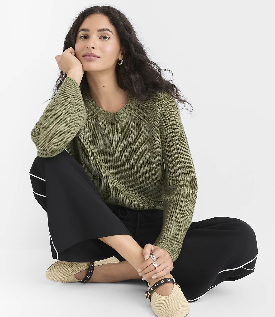 A woman in a green sweater and black pants sits cross-legged against a plain white background.