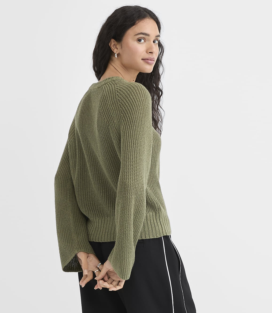 A woman in a green sweater and black pants stands against a plain white background.