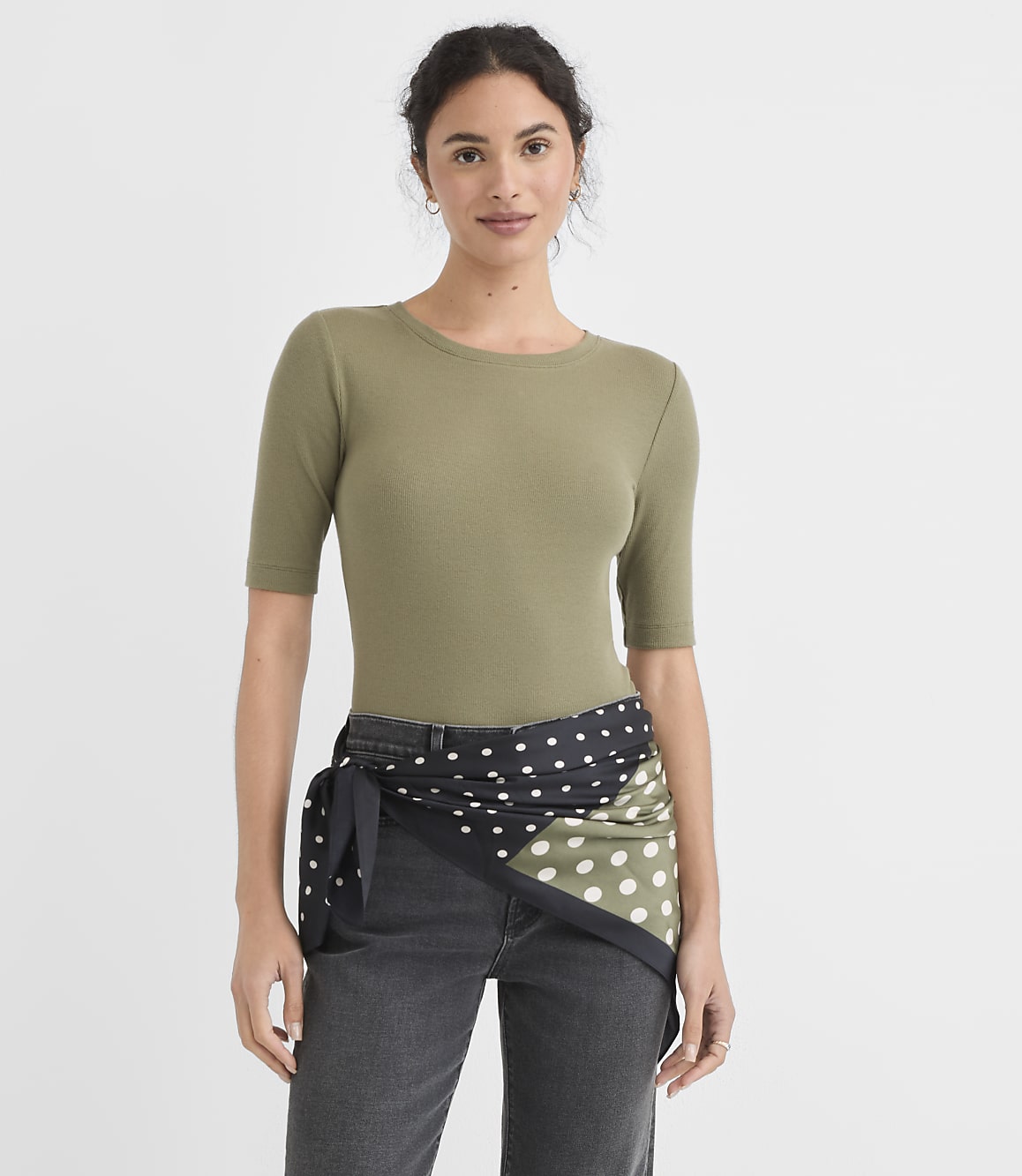 A woman in a green ribbed top and dark jeans stands against a plain white background, with a polka-dotted scarf tied around her waist.
