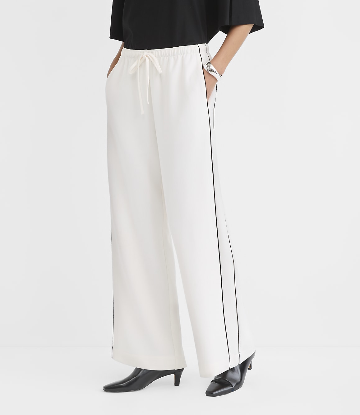 A person stands in white wide-leg pants with black piping and black pointed-toe heels against a plain white background.