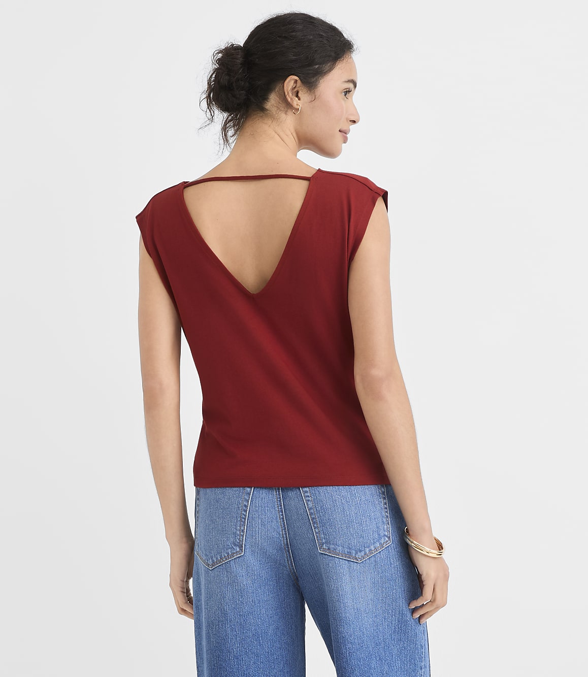 A woman in a maroon v-neck shirt and blue jeans stands against a plain white background.