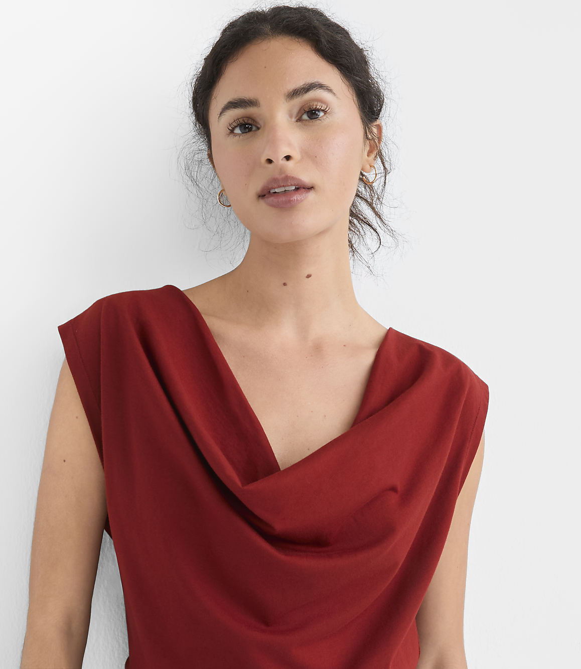 A woman with dark hair and brown eyes, wearing a rust-colored cowl-neck top, is shown against a plain white background.