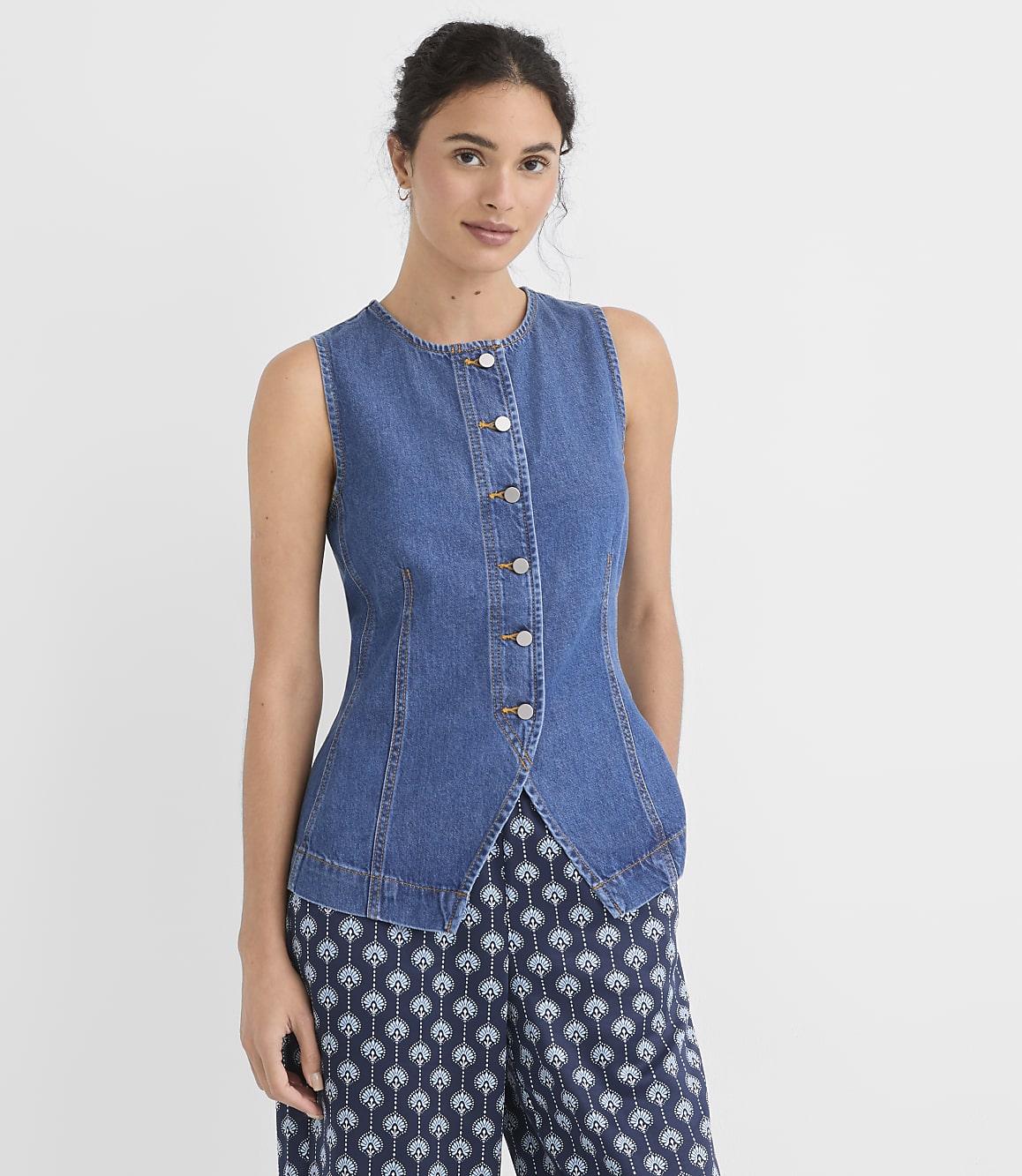 A woman in a denim vest and patterned pants stands against a plain white background.