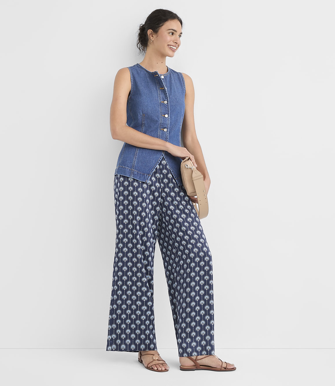 A woman in a denim vest and patterned pants stands against a plain white background, holding a tan clutch.