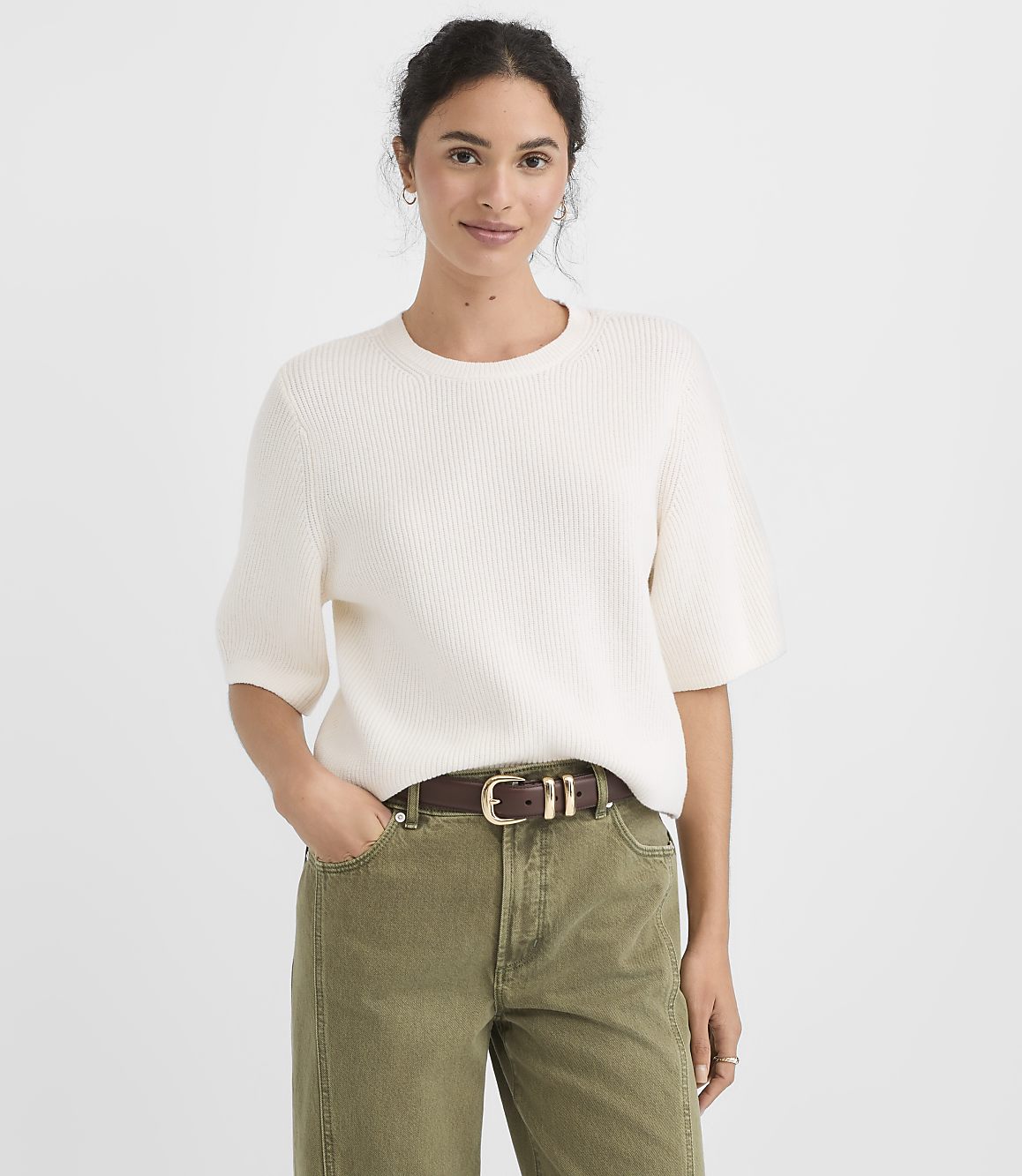 A woman in a cream sweater and olive pants stands against a plain white background.