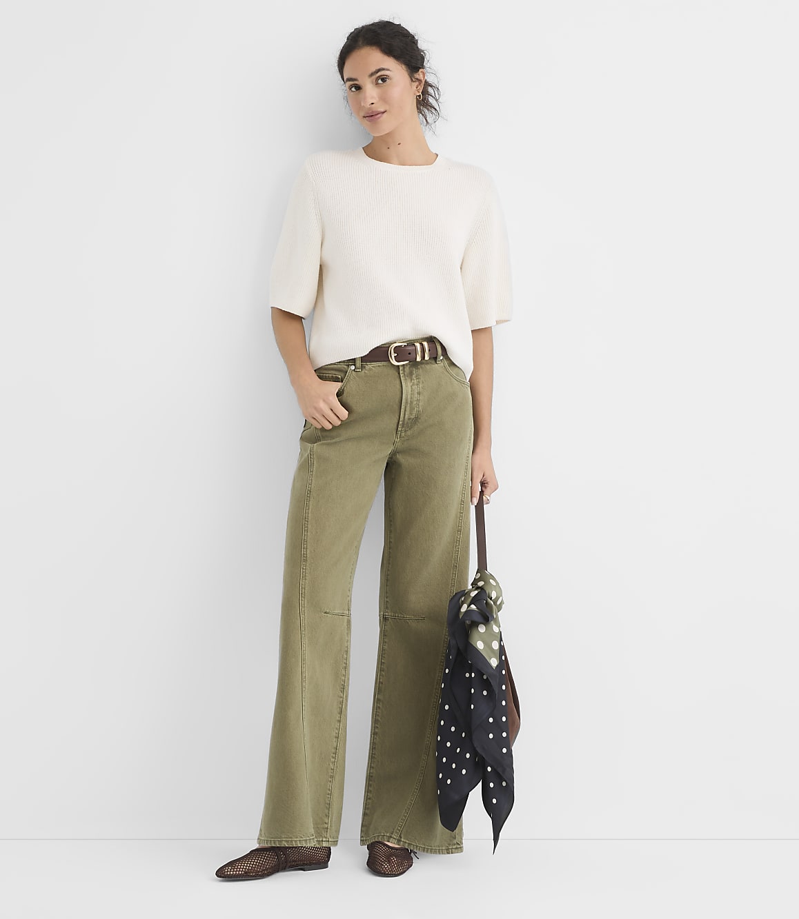 A woman in a cream sweater and olive green wide-leg pants stands against a plain white background, holding a patterned scarf.