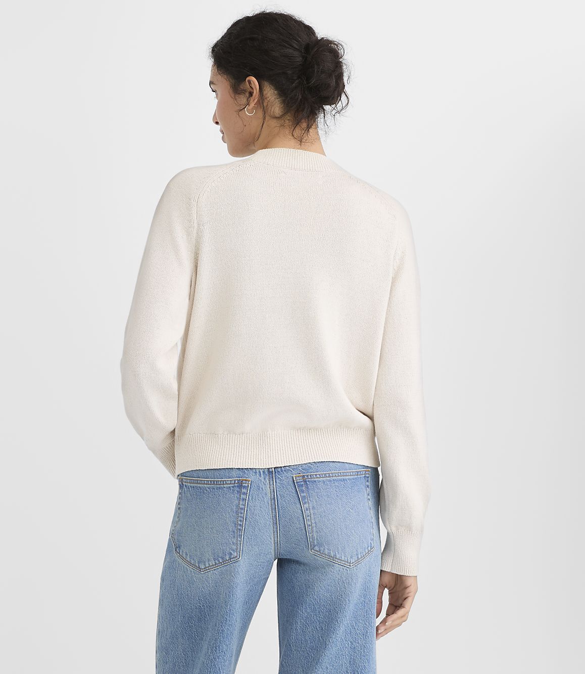 A woman in a cream sweater and blue jeans stands against a plain white background.