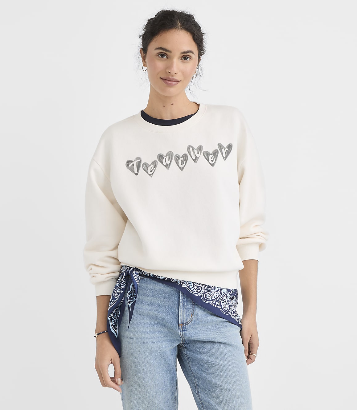A woman in a cream sweatshirt with heart graphics and a blue bandana tied around her waist stands against a plain white background.