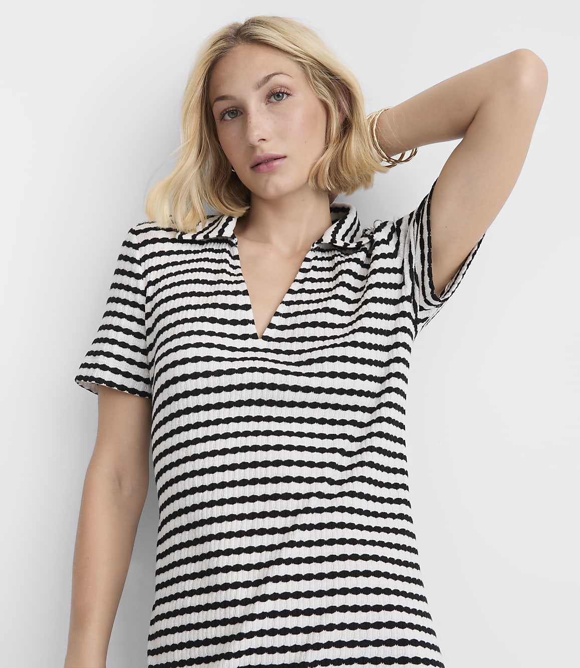 A blonde woman in a striped shirt poses with her arm behind her head against a plain white background.