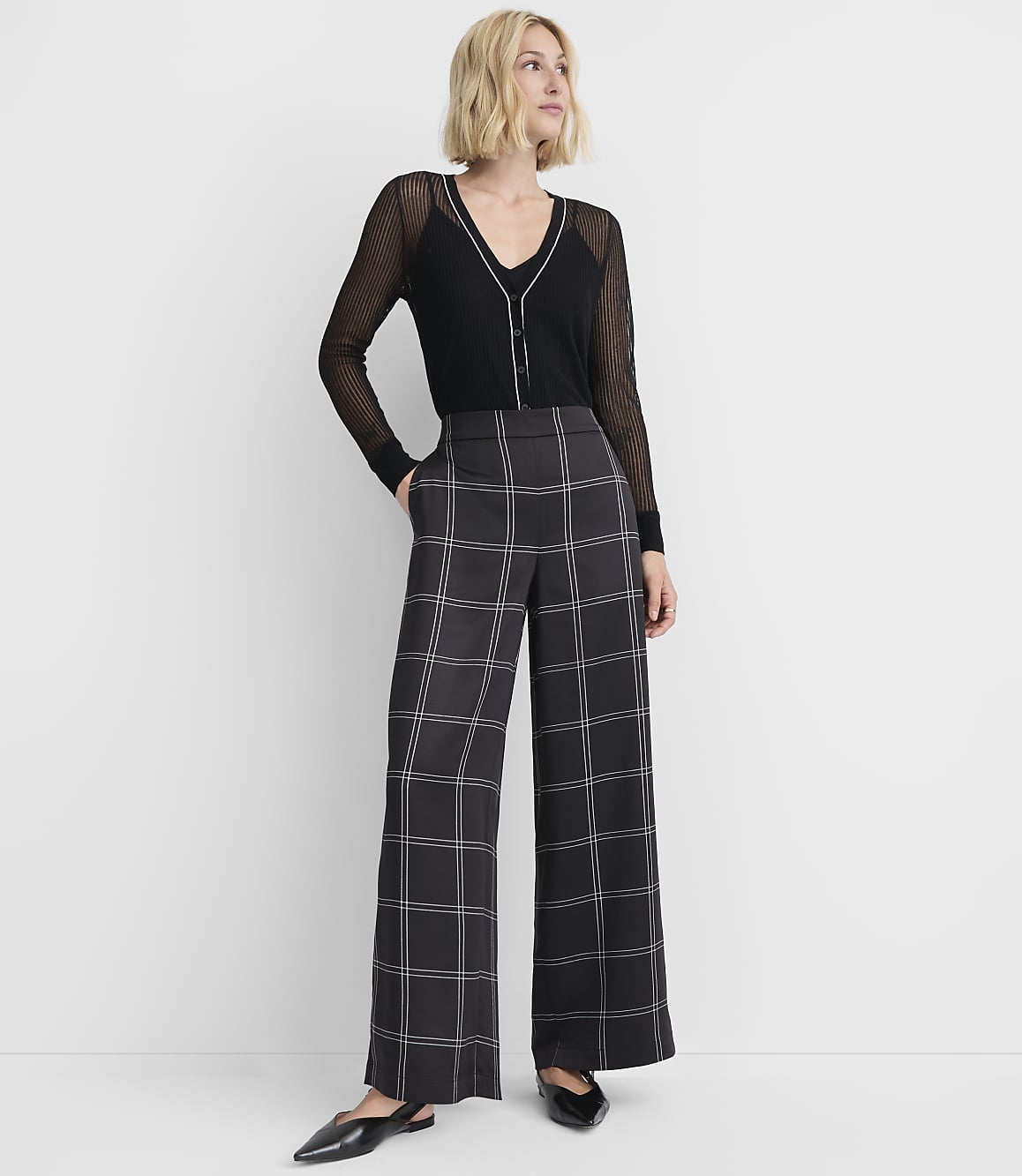A woman in a black sheer top and wide-leg plaid pants stands against a plain white background.